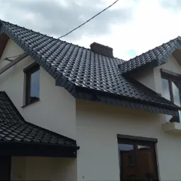 Dom jednorodzinny z nowym, ciemnoszarym dachem z dachówki karpiówki, widoczne okna dachowe i komin z cegły, jasna elewacja budynku, pochmurne niebo w tle.