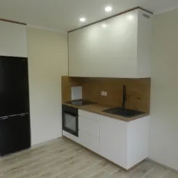 Nowoczesna, minimalistyczna kuchnia z białymi szafkami, drewnianym blatem i czarnym zlewem oraz lodówką, w jasnym pomieszczeniu z płytkami imitującymi drewno na podłodze.