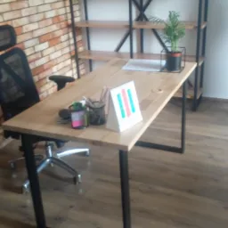 Drewniany blat biurka z czarnymi, metalowymi nogami, zorganizowane miejsce pracy z przybornikami i roślinką w tle ceglana ściana i regał z drewnianymi półkami na metalowym stelażu.