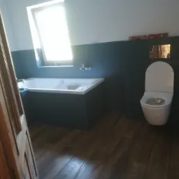 Wnętrze łazienki w trakcie prac wykończeniowych: zabudowana wanna z białą obudową, podwieszana toaleta, ciemne płytki na ścianach do połowy wysokości, drewnopodobne panele na podłodze, poziomica...