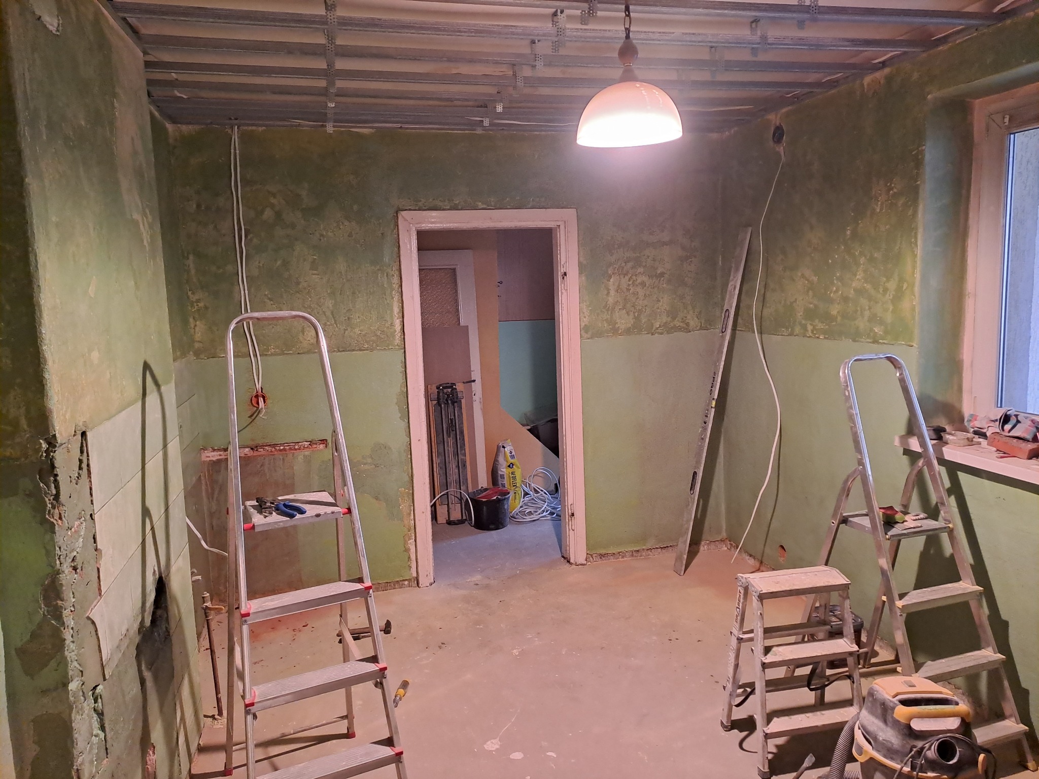 Pokój w trakcie prac przygotowawczych do remontu: odsłonięte instalacje, drabiny, narzędzia, widoczne ślady po starych płytkach na ścianach, konstrukcja sufitu podwieszanego