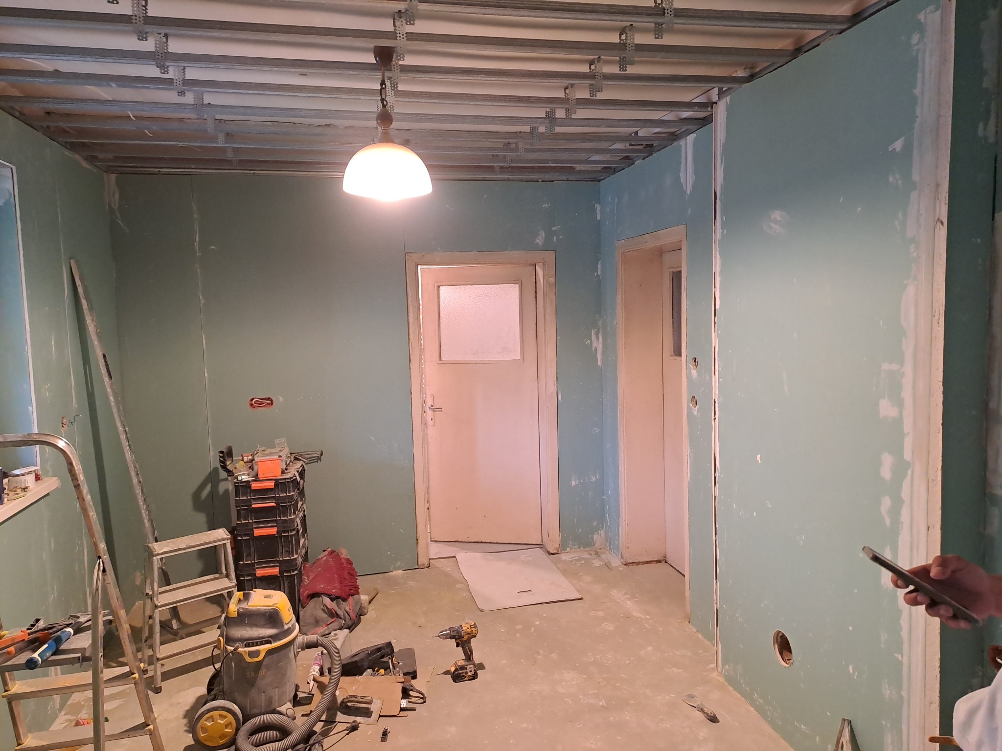 Wnętrze w trakcie prac w Pile: widoczne ściany wyłożone płytami kartonowo-gipsowymi, metalowa konstrukcja sufitu podwieszanego, narzędzia i odkurzacz budowlany.