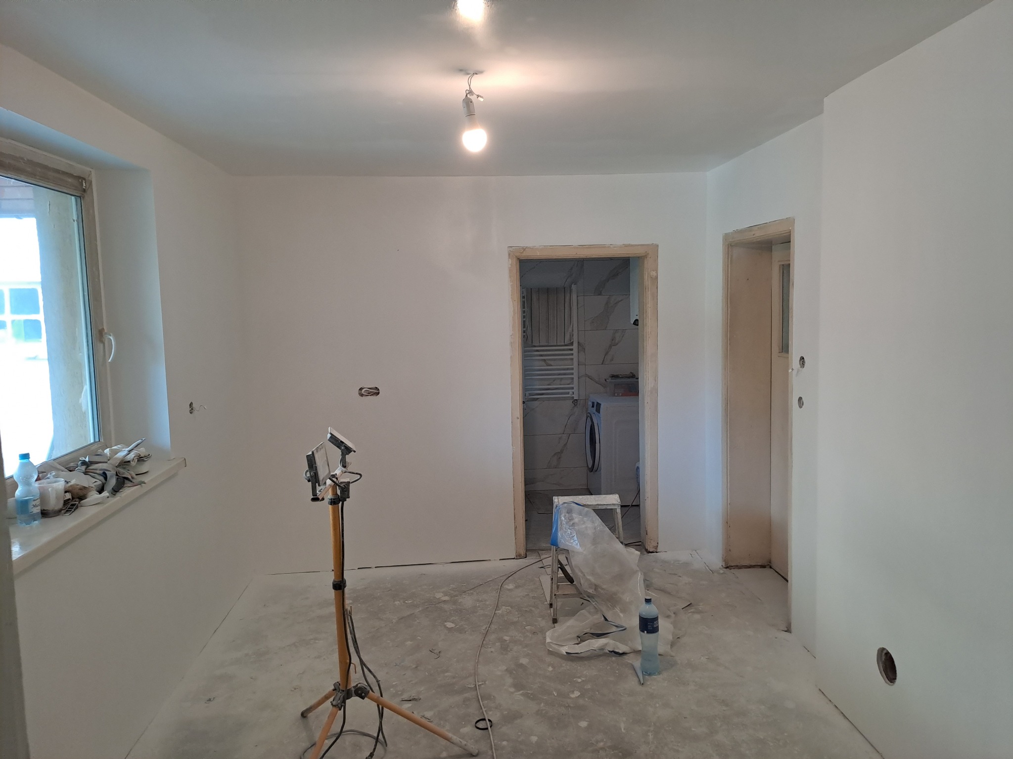 Wnętrze w trakcie malowania na biało; widoczna lampa budowlana na statywie, okno z narzędziami na parapecie, wejście do łazienki z widoczną pralką i grzejnikiem oraz drugie wejście do innego...