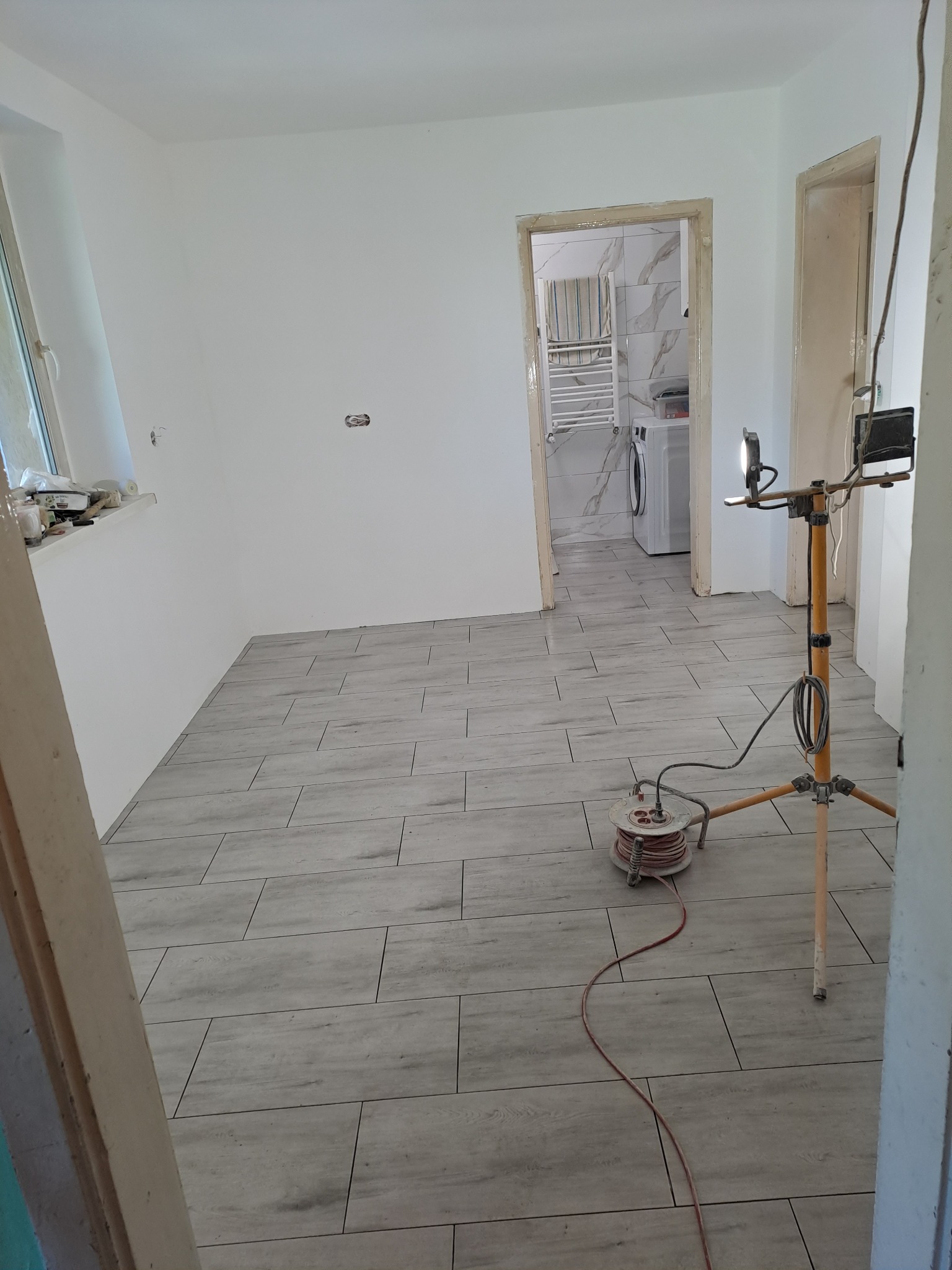 Wnętrze w trakcie prac wykończeniowych: ułożona szara podłoga imitująca drewno, widoczna łazienka z białymi płytkami i pralką w tle, lampa budowlana na trójnogu z przedłużaczem.
