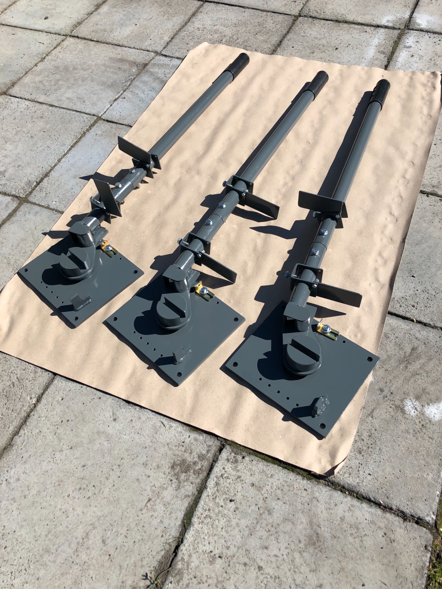 Trzy szare, regulowane podpory teleskopowe z kwadratowymi podstawami, leżące na brązowym papierze na betonowym podłożu.