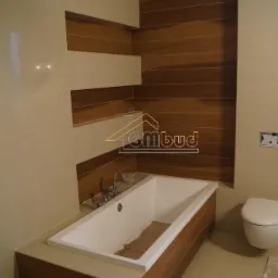 Wykończona łazienka z wanną obudowaną brązowymi panelami imitującymi drewno, jasne płytki na ścianach i podłodze, widoczne logo firmy GMbud.