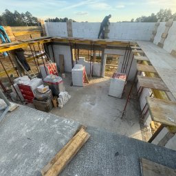 GMBUD pl - budowa DOMów - poddasza - Grzegorz Motyka Dąbrowa Tarnowska - Wnętrze budowanego domu z bloczków, widok z góry na szalunki stropowe, materiały budowlane i dwóch robotników przy wznoszeniu ścian.