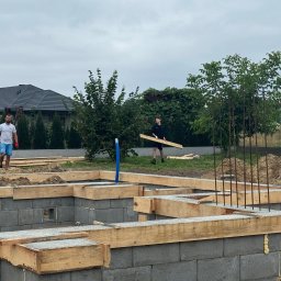 GMBUD pl - budowa DOMów - poddasza - Grzegorz Motyka Dąbrowa Tarnowska - Wykonywanie fundamentów pod budynek z bloczków betonowych, drewniane szalunki i wystające pręty zbrojeniowe, dwóch mężczyzn na placu budowy, widok na dom w tle