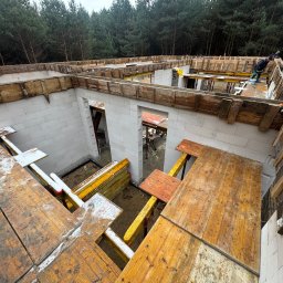 GMBUD pl - budowa DOMów - poddasza - Grzegorz Motyka Dąbrowa Tarnowska - Wznoszenie ścian domu z bloczków, widoczne drewniane szalunki stropowe i pracownicy na górze, tło lasu.