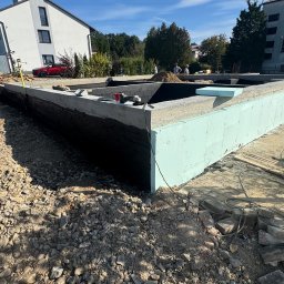 GMbud - budowa DOMów - poddasza - Grzegorz Motyka Dąbrowa Tarnowska - Ocieplone fundamenty domu jednorodzinnego w trakcie budowy, widoczne narzędzia pracy i fragment budynku w tle. Izolacja pionowa i termiczna.