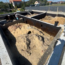 GMbud - budowa DOMów - poddasza - Grzegorz Motyka Dąbrowa Tarnowska - Fundamenty z izolacją bitumiczną w Dąbrowie Tarnowskiej. Wnętrze wypełnione piaskiem, widoczni pracownicy budowlani. Ujęcie z góry, w tle domy jednorodzinne.