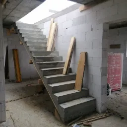 Surowe betonowe schody wewnętrzne w trakcie budowy w domu z bloczków betonowych, z drewnianymi deskami opartymi o ścianę i schody, widoczny materiał izolacyjny Termobet.