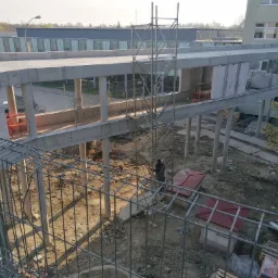 Budowa betonowego łącznika między budynkami, widok z góry przez metalowe ogrodzenie, widoczne rusztowania i pracownik na drabinie, w tle budynek z oknami.