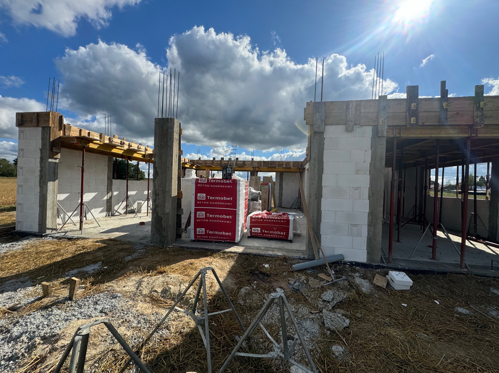 Wznoszenie ścian parterowego domu z betonu komórkowego Termobet, widoczne zbrojenia pionowe, podpory szalunków i palety z materiałem, w tle niebo z chmurami.