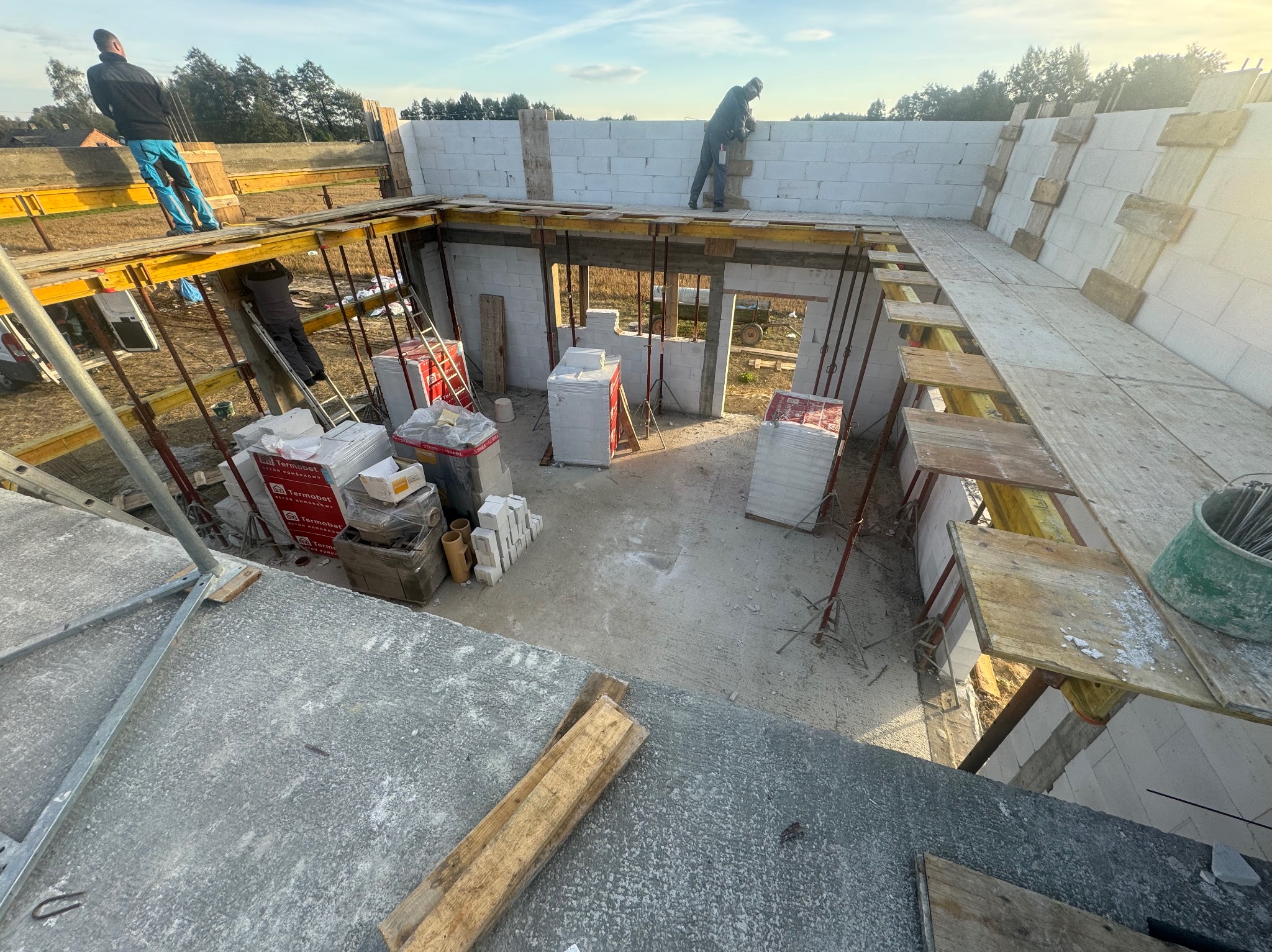 Wnętrze budowanego domu z bloczków, widok z góry na szalunki stropowe, materiały budowlane i dwóch robotników przy wznoszeniu ścian.