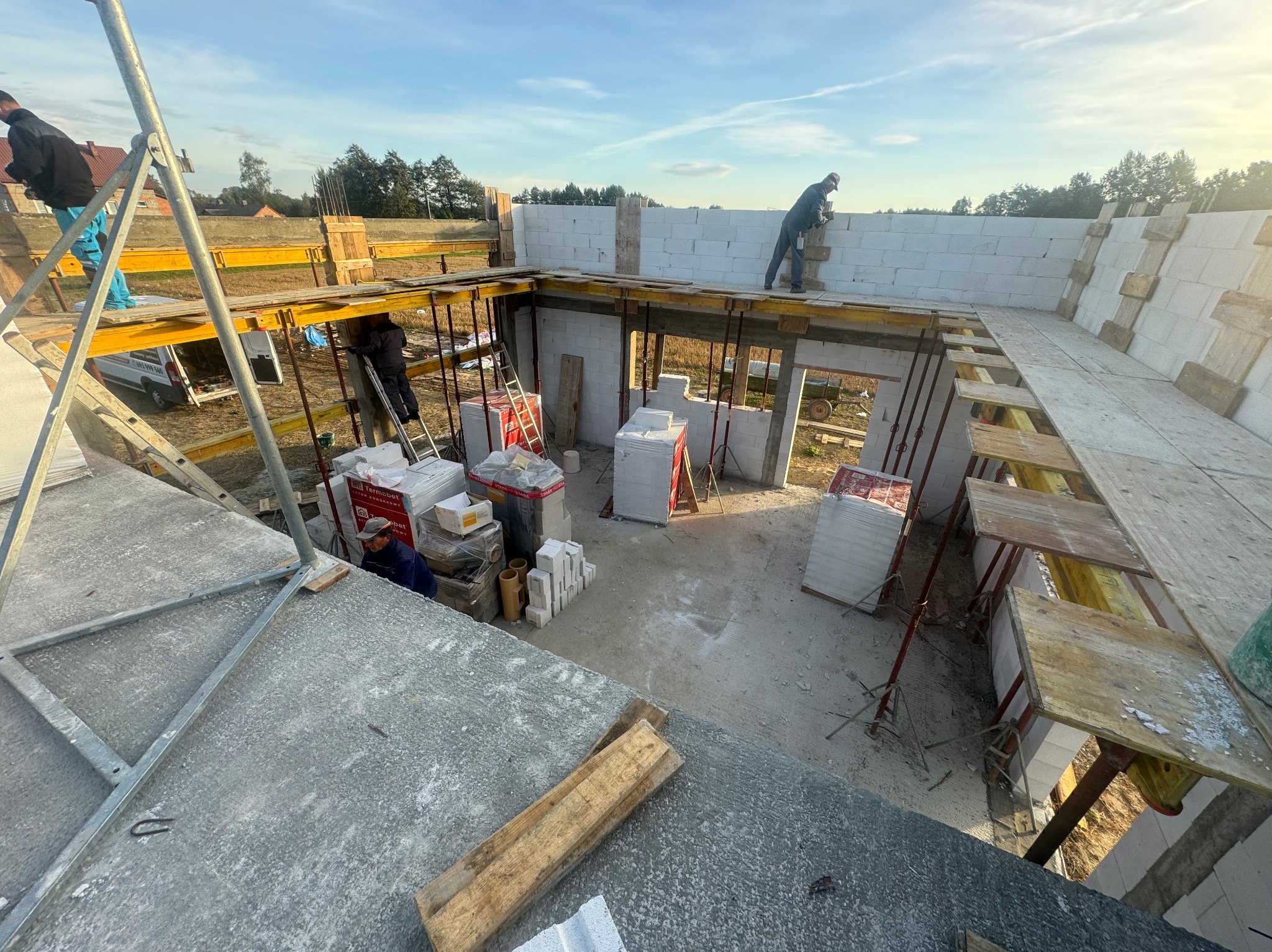 Budowa domu z białych bloczków: widok z góry na częściowo wzniesione ściany, drewniane szalunki, stosy materiałów budowlanych i pracujących robotników w porannym świetle.