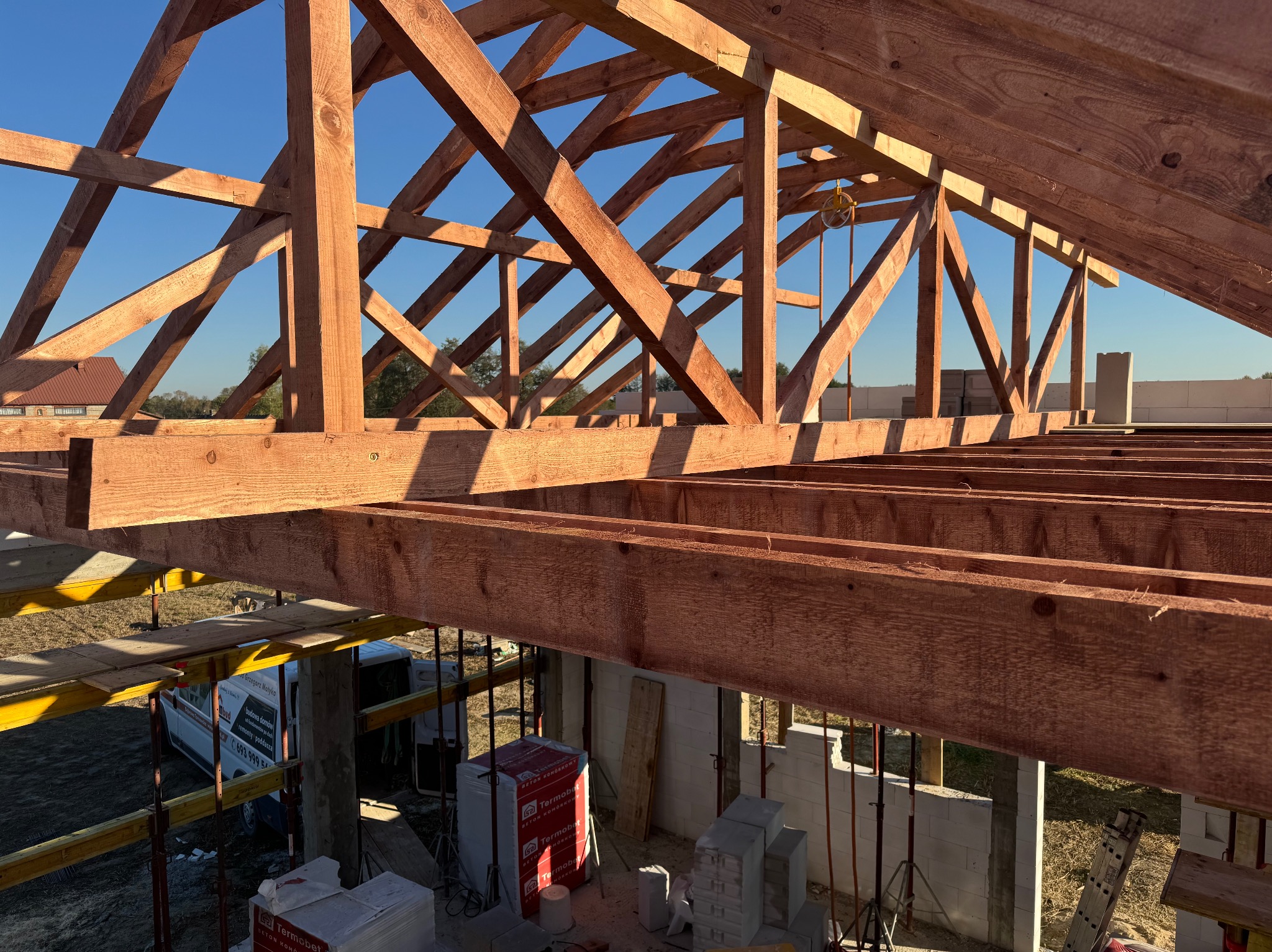 Konstrukcja drewniana dachu w trakcie budowy, widok od wewnątrz, z widocznymi elementami szalunku i pustakami ceramicznymi oraz brygadą budowlaną w tle.