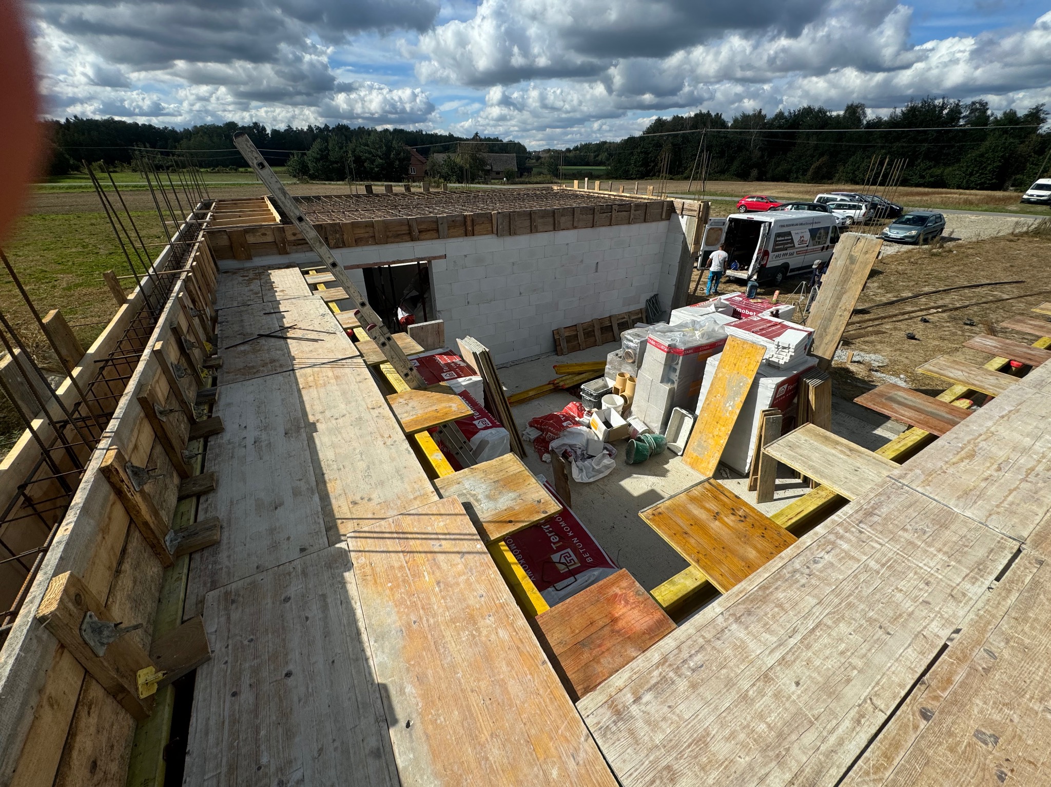 Budowa domu murowanego w trakcie: widok z góry na fundamenty, ściany z bloczków i drewniane szalunki, z zaparkowanym samochodem dostawczym firmy w tle.