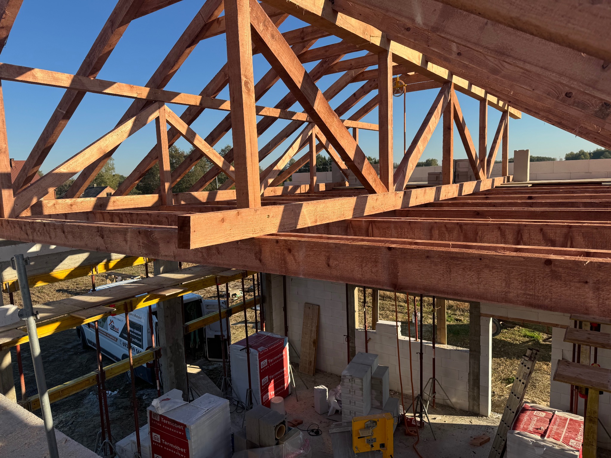 Wnętrze budowy domu w stanie surowym otwartym, widoczna drewniana konstrukcja dachu, ściany z betonu komórkowego, materiały budowlane na placu i samochód dostawczy na zewnątrz.