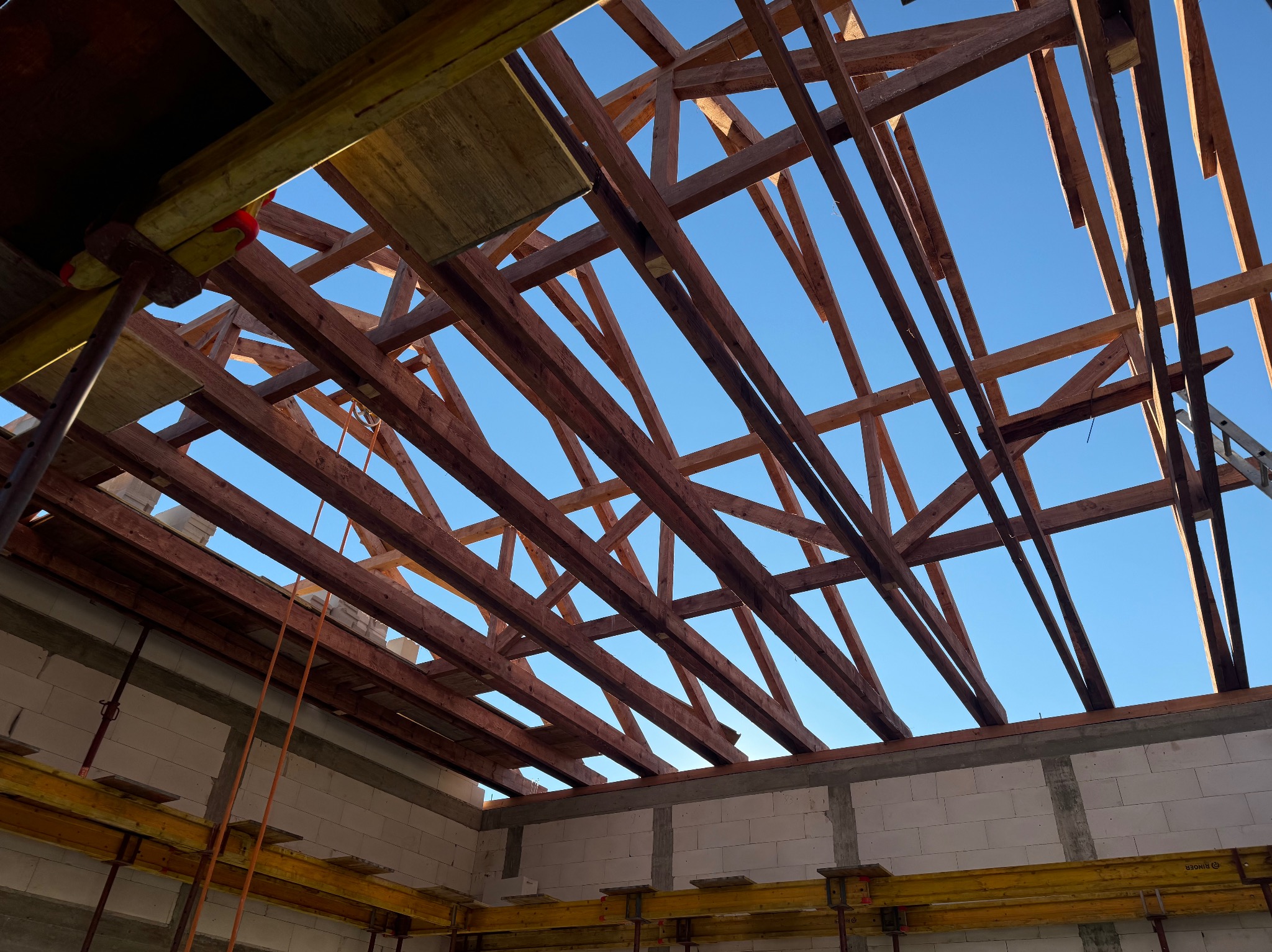 Konstrukcja więźby dachowej z drewnianych belek na tle błękitnego nieba, widok z wnętrza budowanego domu z białych bloczków.