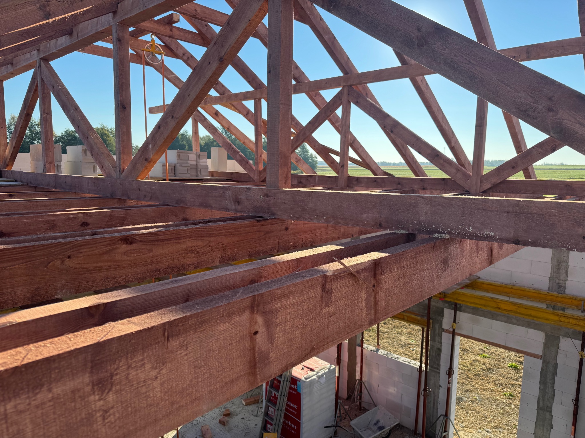 Wnętrze budowanego domu z widoczną drewnianą konstrukcją dachu i fragmentem muru z bloczków, zarys zielonego pola w tle pod błękitnym niebem.