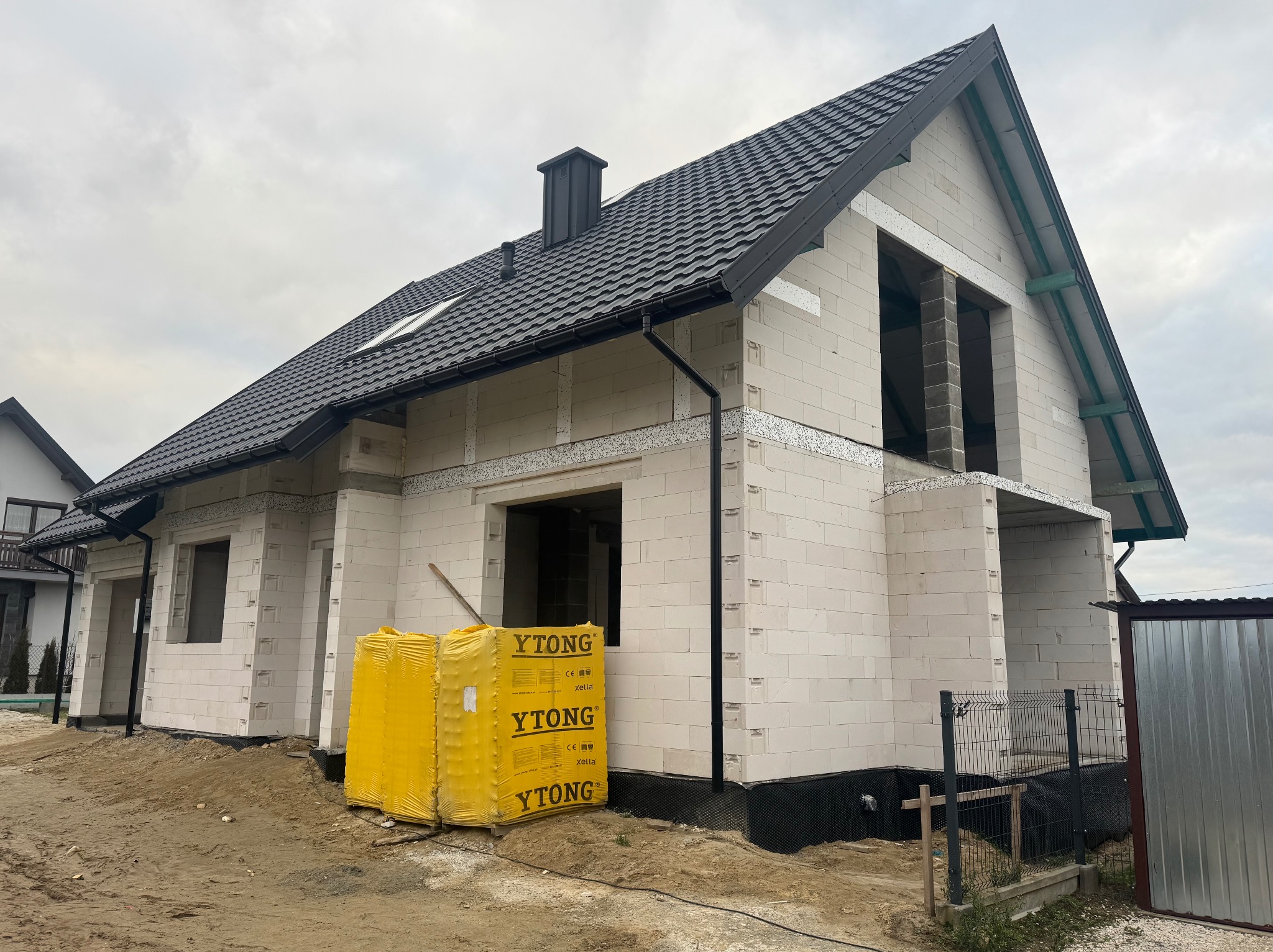 Surowy stan domu z białych bloczków Ytong, z czarną dachówką i orynnowaniem, widoczne żółte palety z bloczkami Ytong, okno dachowe, komin i fundament zabezpieczony folią.