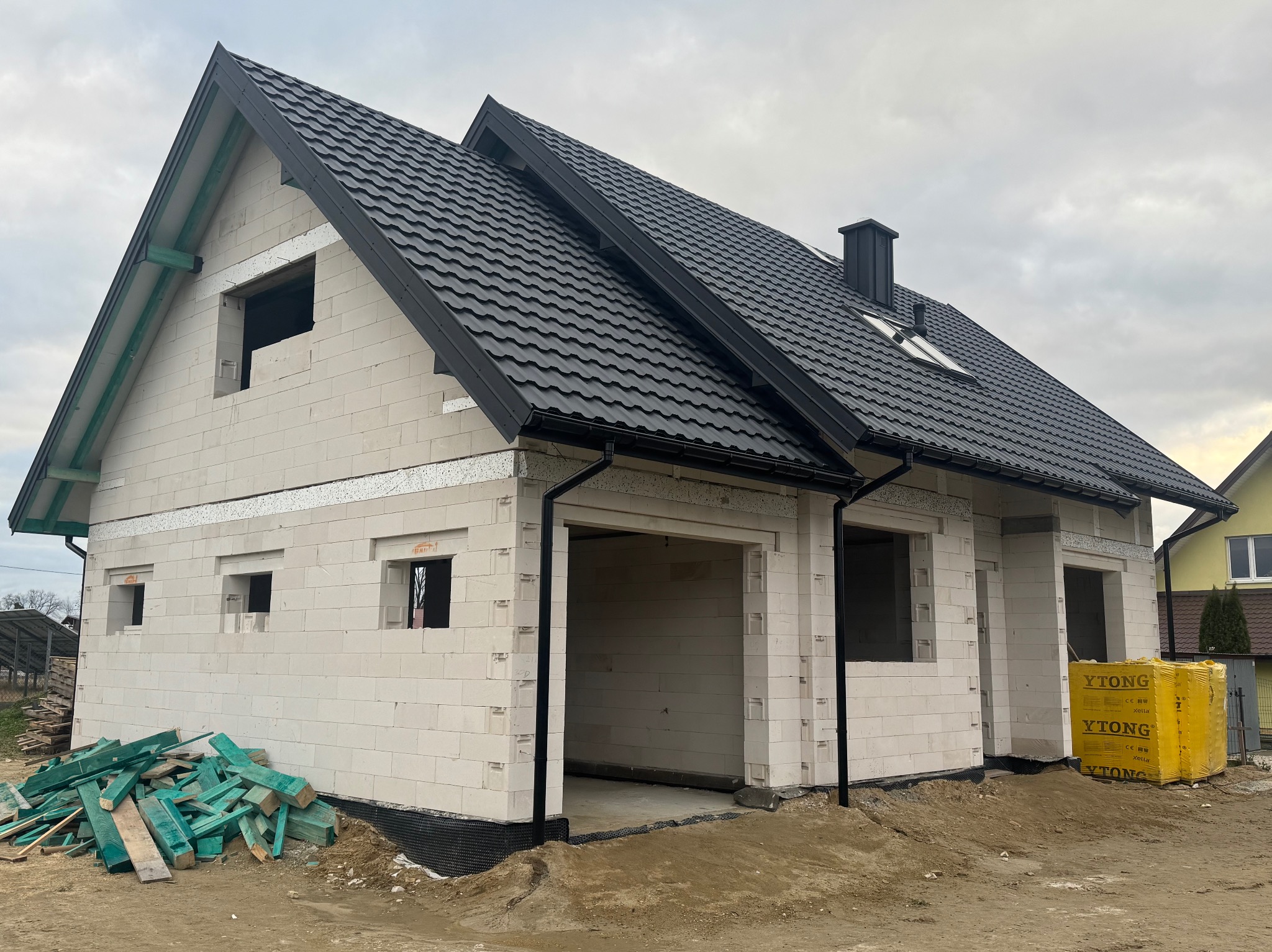 Niedokończony dom murowany z czarną dachówką, widoczne bloczki Ytong i składowane drewno budowlane przy fundamencie.