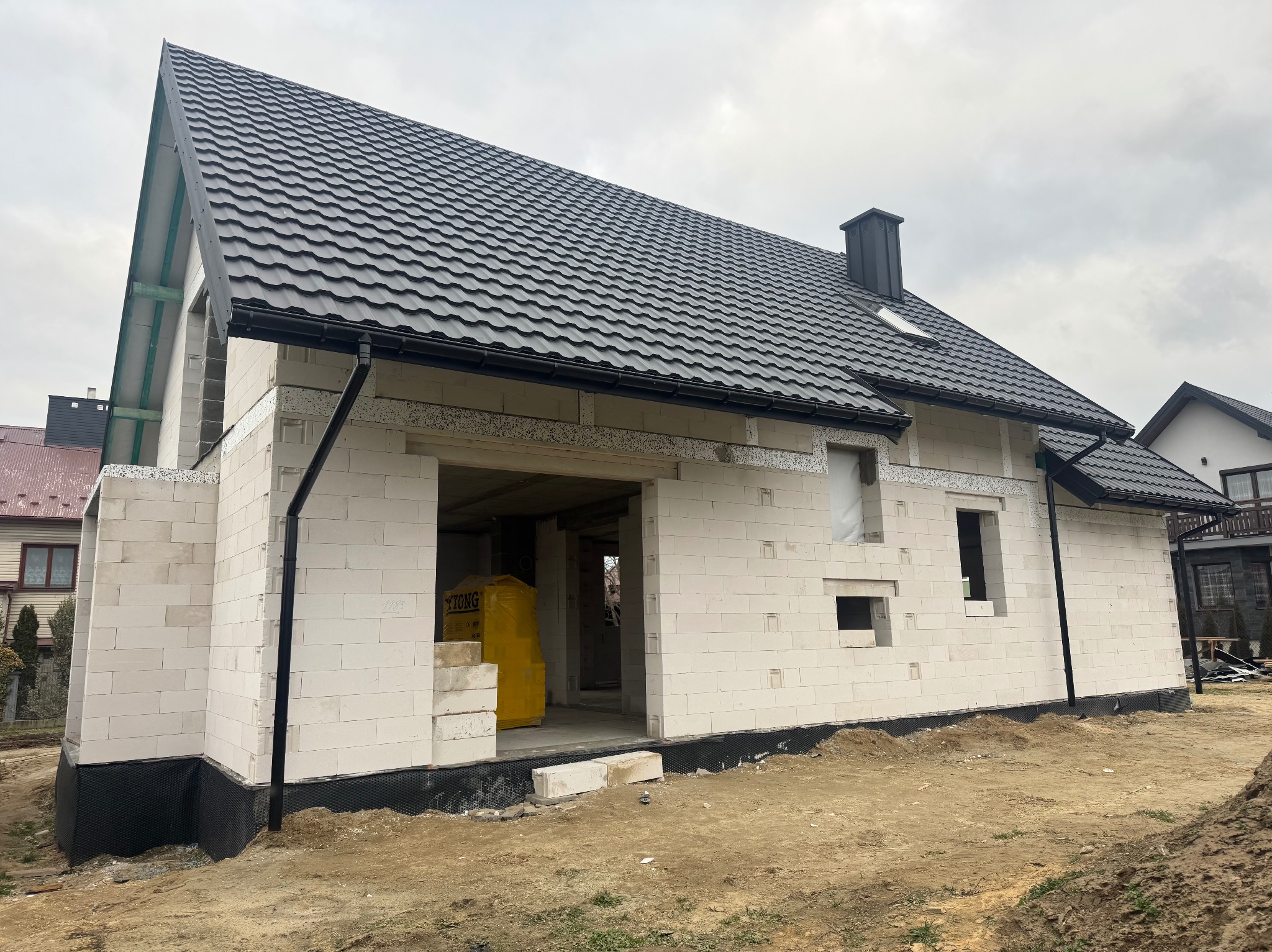 Surowy stan domu z białego betonu komórkowego z czarnym dachem, widoczne przygotowane otwory okienne i drzwiowe, z żółtymi paletami materiałów wewnątrz, na tle pochmurnego nieba.