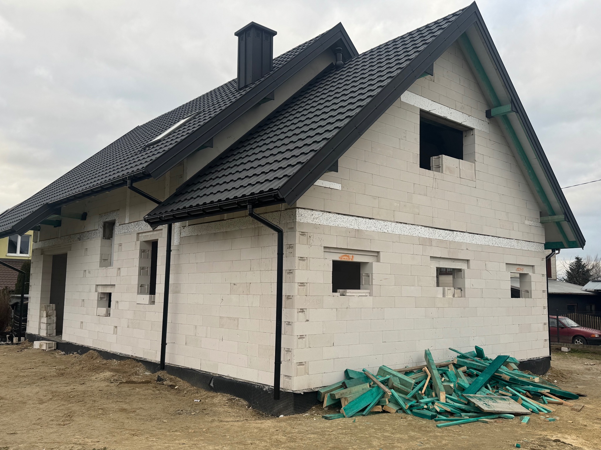 Surowy stan domu z białych bloczków z ciemnym dachem, widoczne otwory okienne i drzwiowe, oraz stos zielonych desek na ziemi.