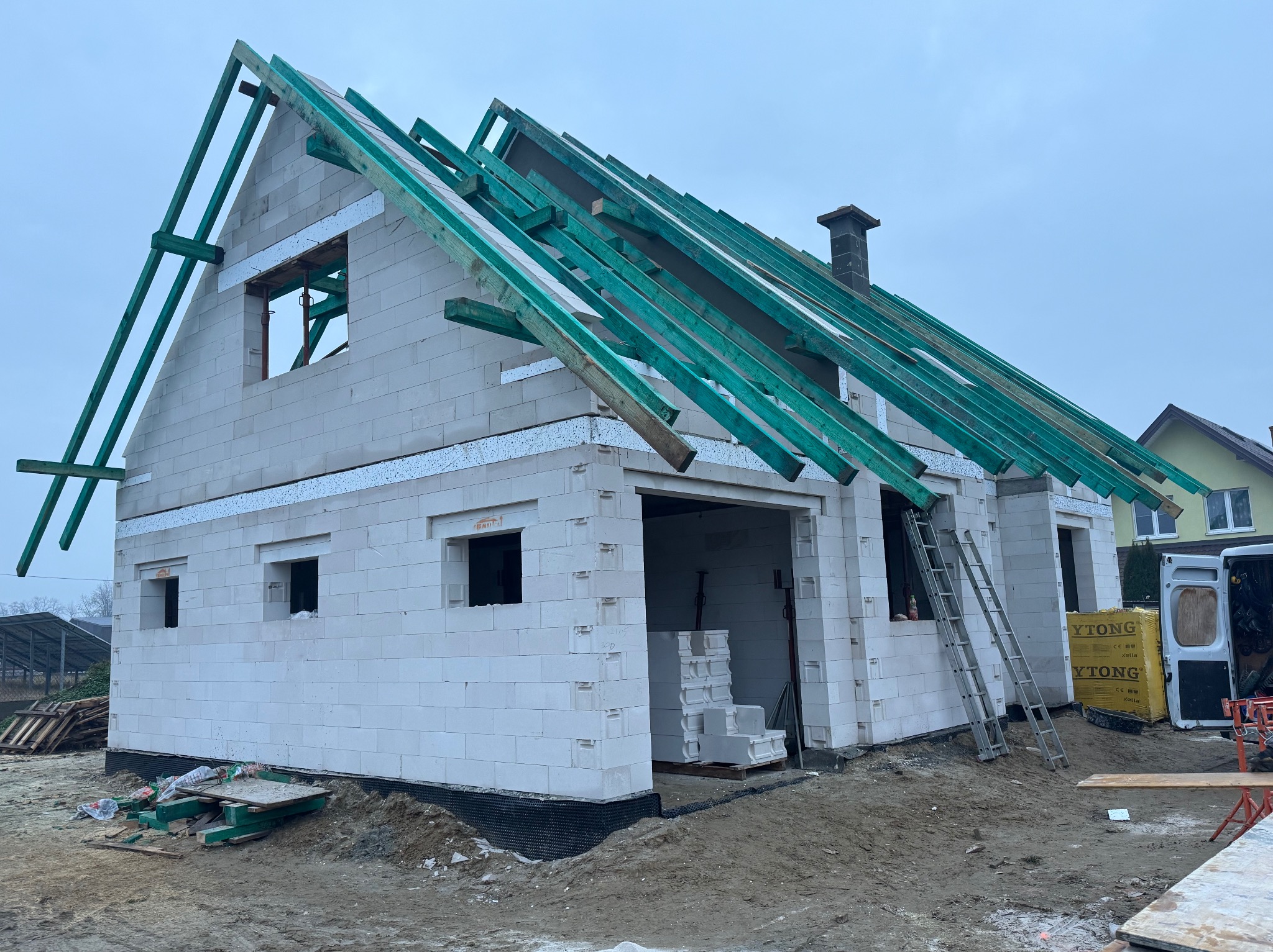 Surowy stan domu z białych bloczków z widoczną konstrukcją dachu z impregnowanego drewna, drabina oparta o ścianę i paleta bloczków Ytong obok furgonetki.