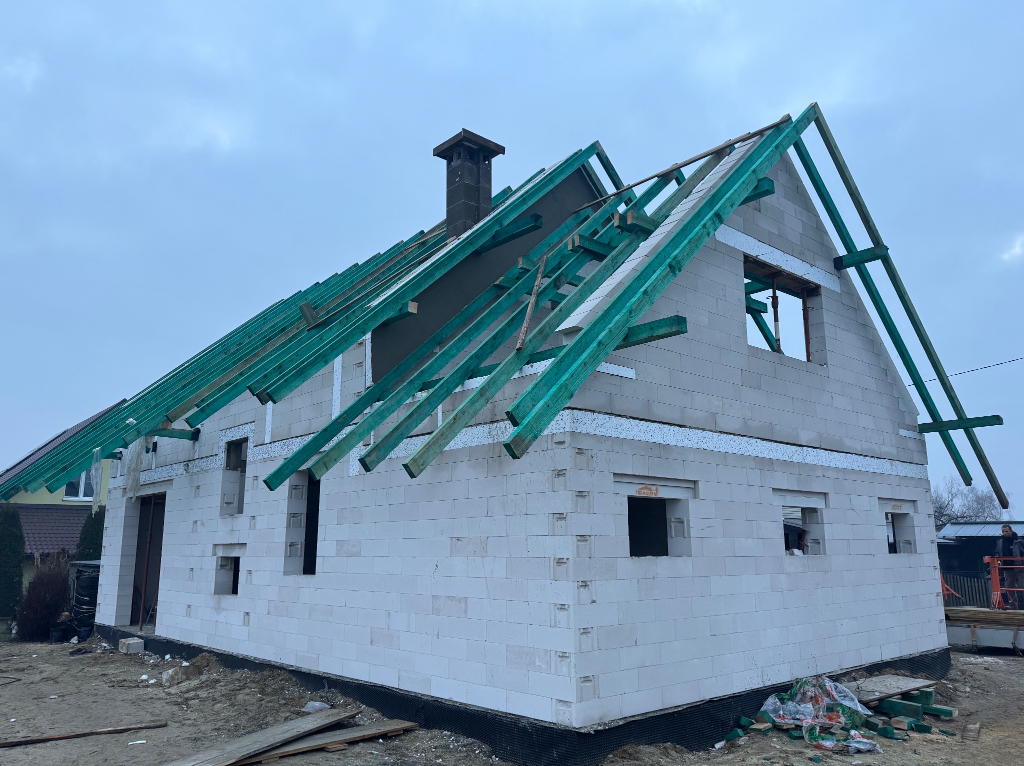 Surowy stan domu z białych bloczków, widoczne zielone krokwie dachu, komin i przygotowane otwory okienne na tle pochmurnego nieba.