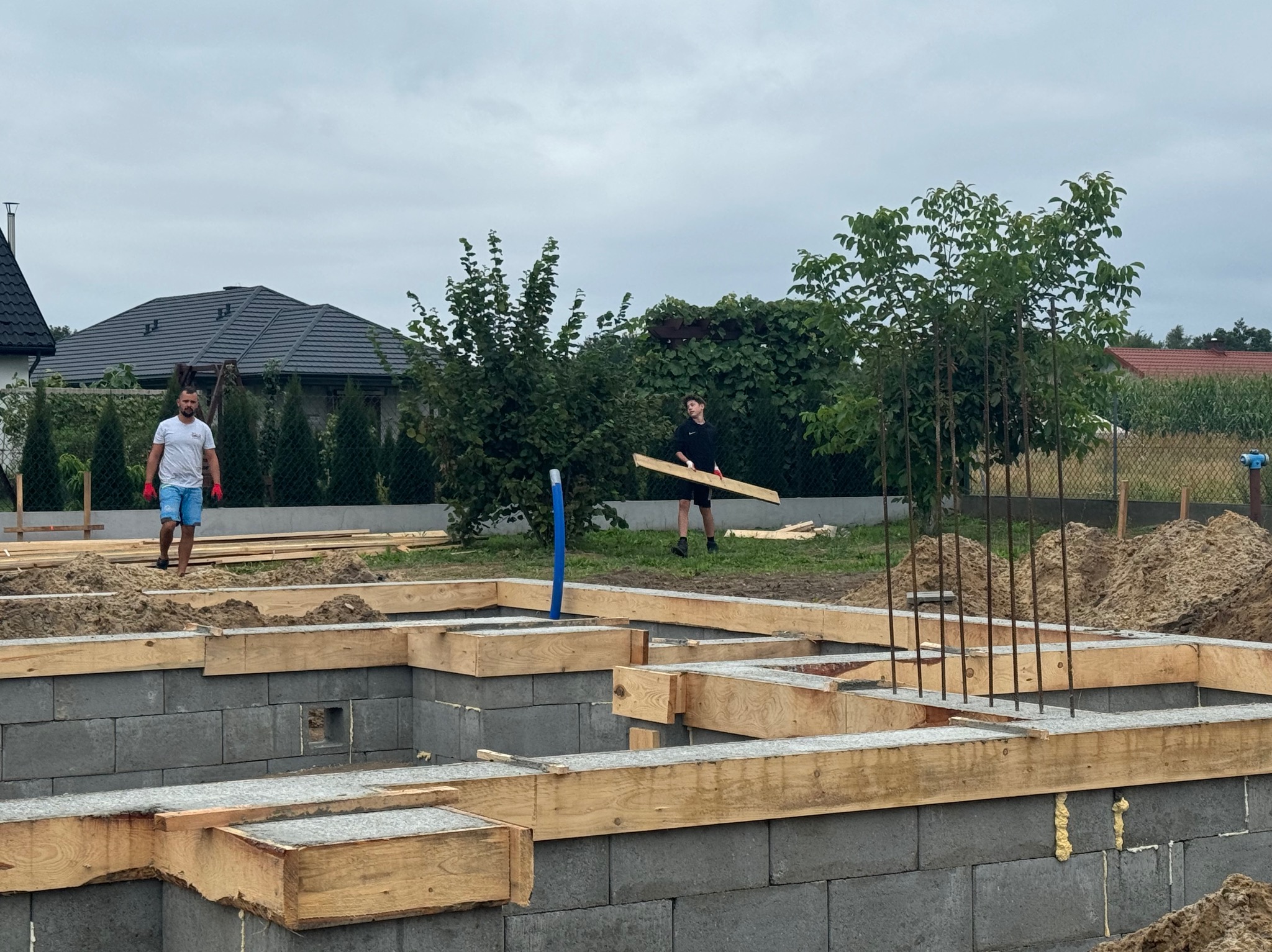 Wykonywanie fundamentów pod budynek z bloczków betonowych, drewniane szalunki i wystające pręty zbrojeniowe, dwóch mężczyzn na placu budowy, widok na dom w tle