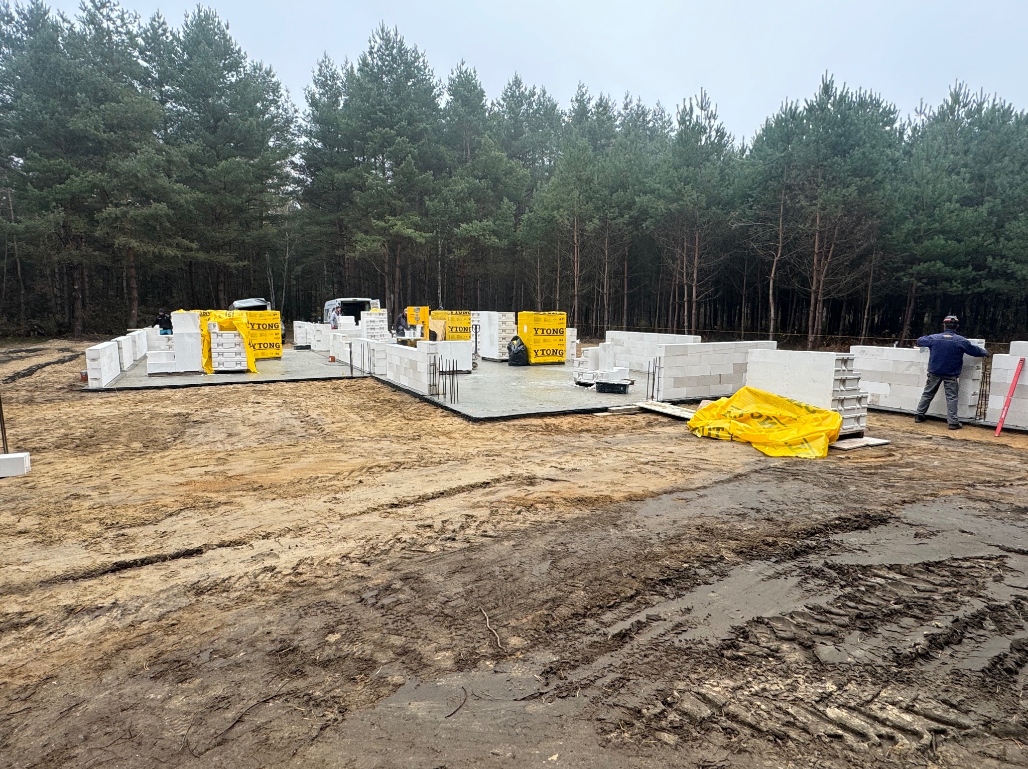 Budowa domu w lesie: fundamenty wylane, pierwsze bloczki Ytong ustawione, teren błotnisty po deszczu, pracownik z łatą murarską przy ścianie.