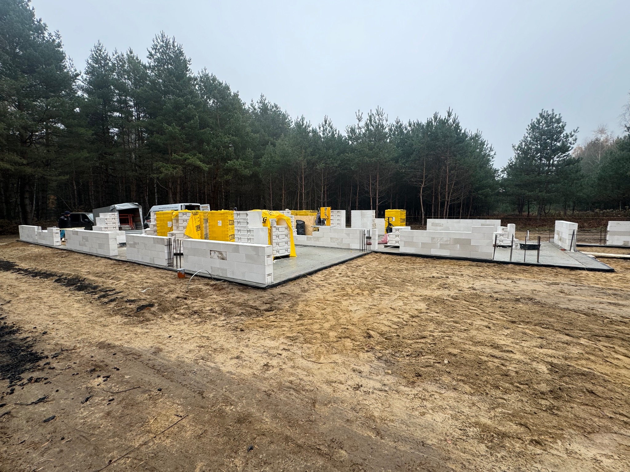 Fundamenty domu w trakcie budowy z bloczków Ytong, widoczne zbrojenia i przygotowane palety bloczków w tle lasu.