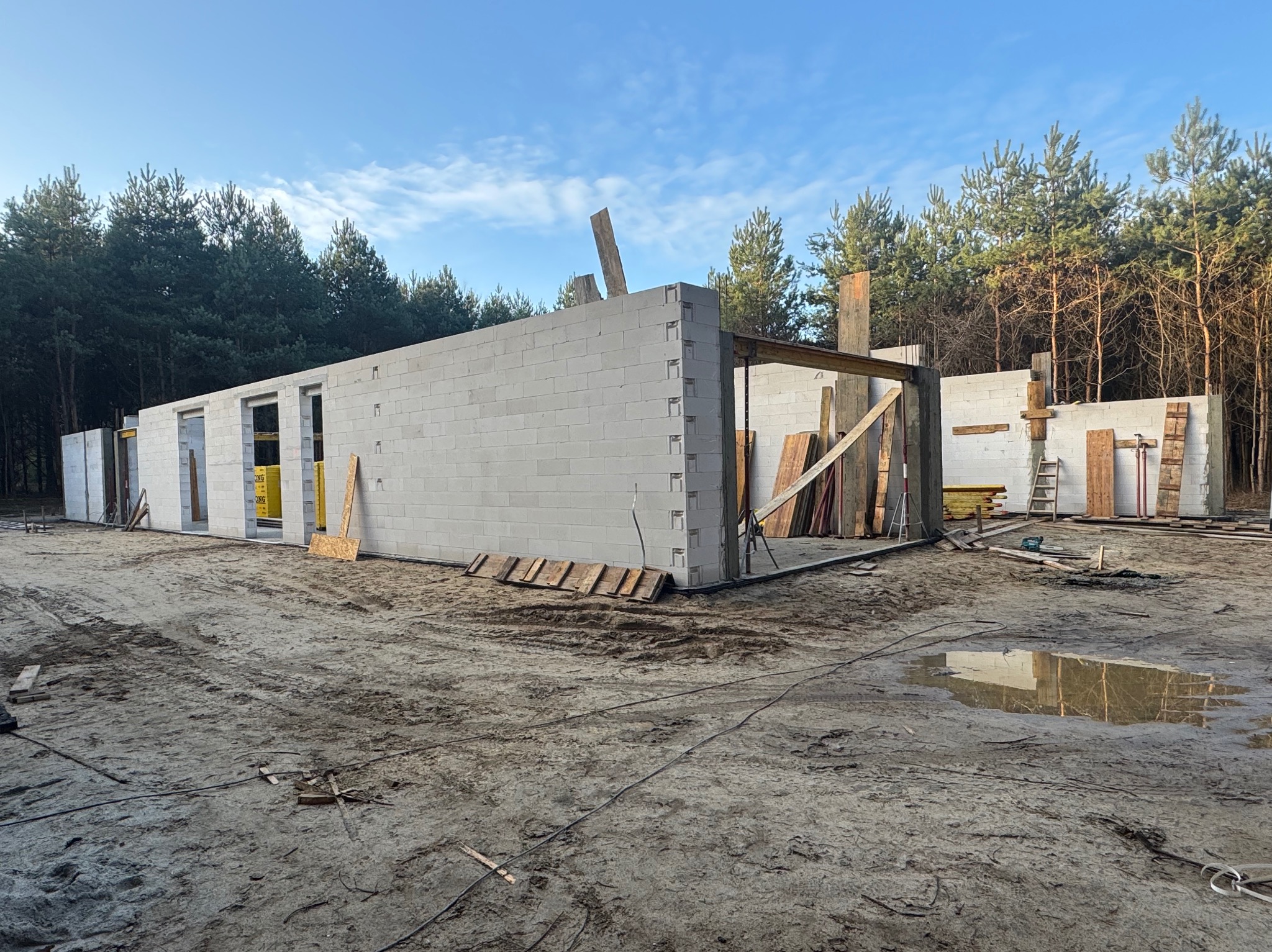 Surowy stan budowy domu z betonu komórkowego, widoczne ściany w trakcie wznoszenia, drewniane podpory i rozległy teren budowy z kałużami po deszczu, las w tle.