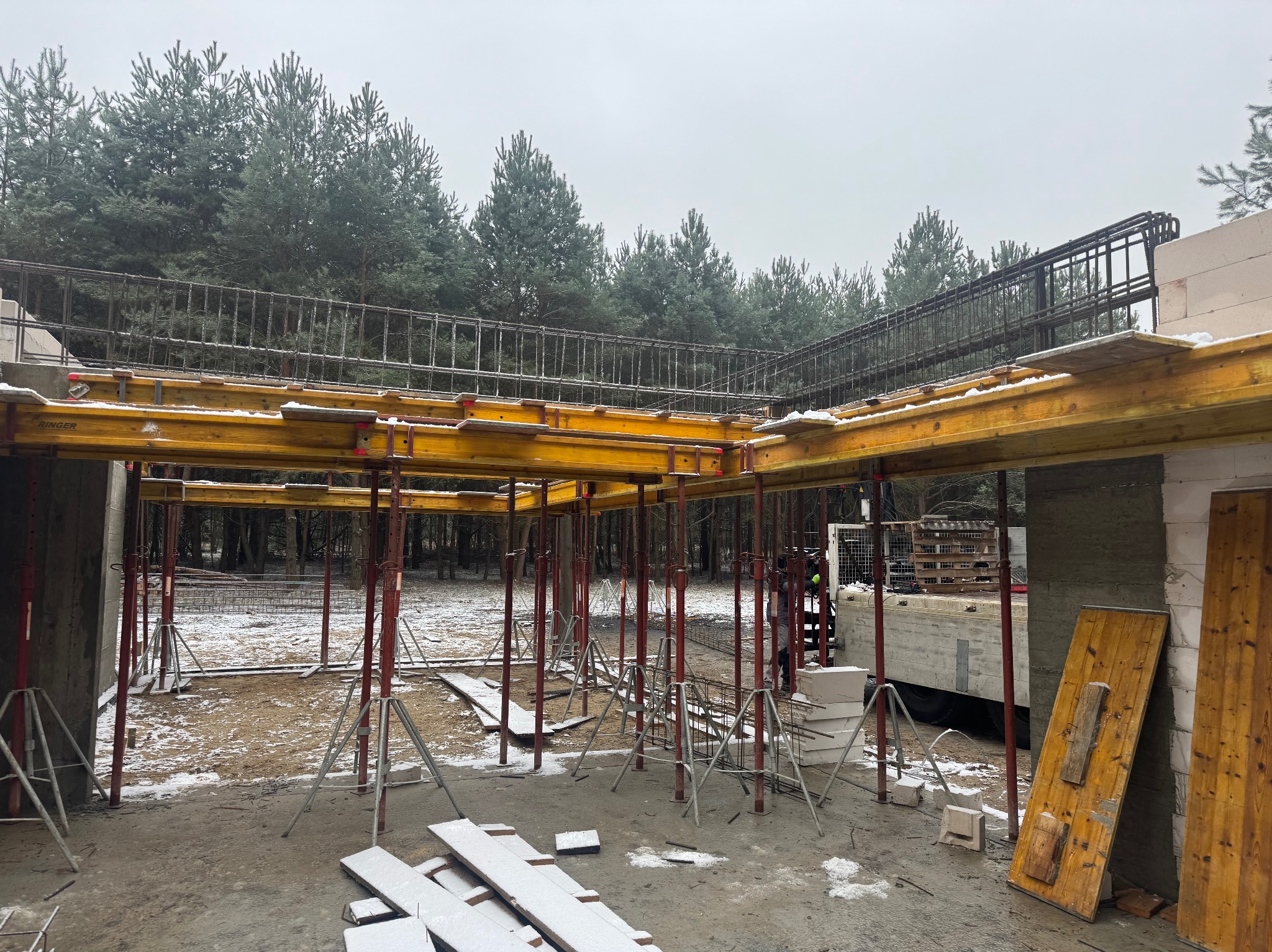 Budowa domu w trakcie zimy: Żółte szalunki podtrzymują zbrojenie stropu, w tle ośnieżone drzewa i częściowo wybudowane ściany z betonu komórkowego.