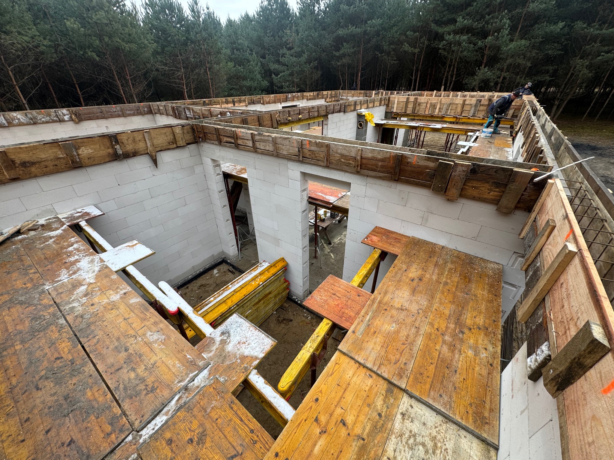 Wznoszenie ścian domu z bloczków, widoczne drewniane szalunki stropowe i pracownicy na górze, tło lasu.