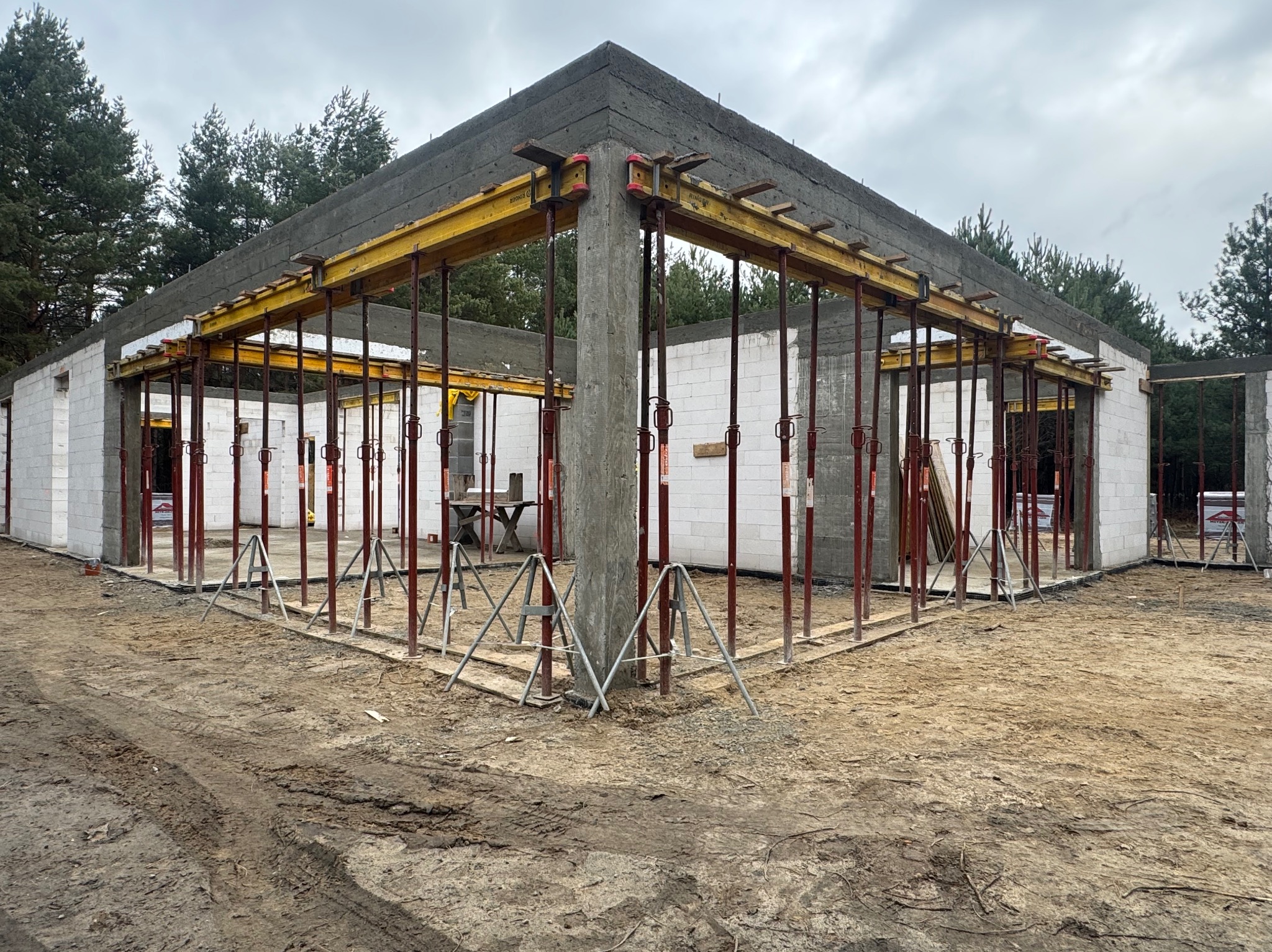 Surowy stan budynku z betonu komórkowego w trakcie budowy z widocznym szalunkiem i podporami, na ziemi ślady opon, w tle drzewa.