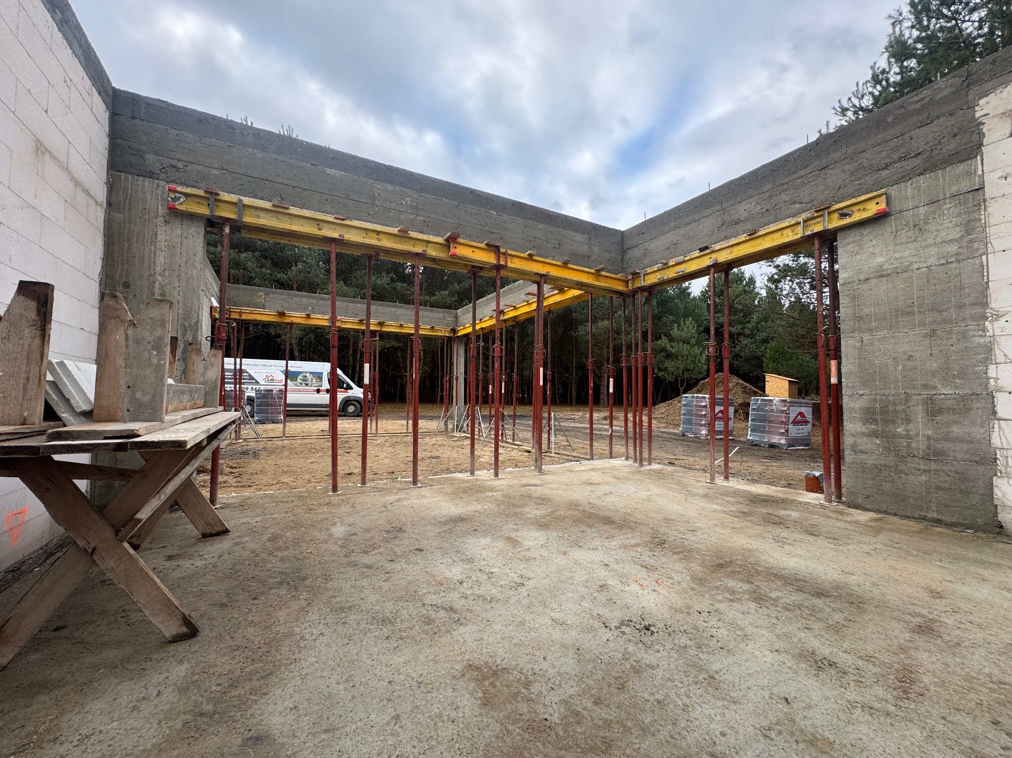 Wnętrze domu w trakcie budowy z widocznymi ścianami z betonu komórkowego i szalunkami stropowymi, na zewnątrz las i zaparkowany biały bus.