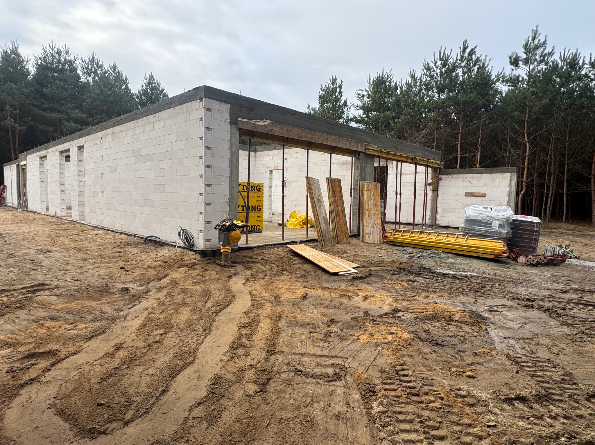 Budowa domu w stanie surowym otwartym z bloczków, widoczne szalunki nad otworami okiennymi i drzwiowymi, teren budowy z błotnistą ziemią i sprzętem budowlanym.