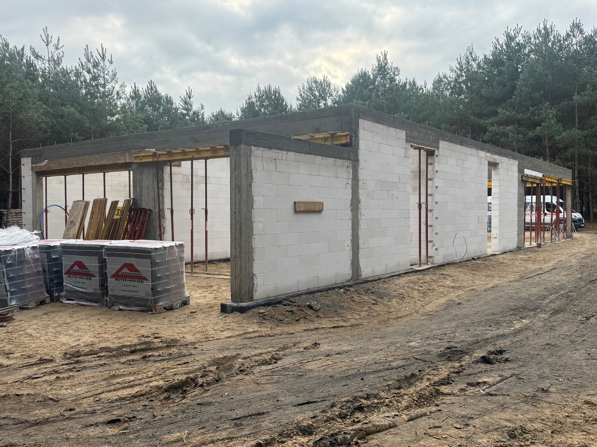 Budowa domu w stanie surowym otwartym z betonu komórkowego, widoczne podpory stropowe, palety z dachówką ceramiczną Meyer-Holsen i las w tle.