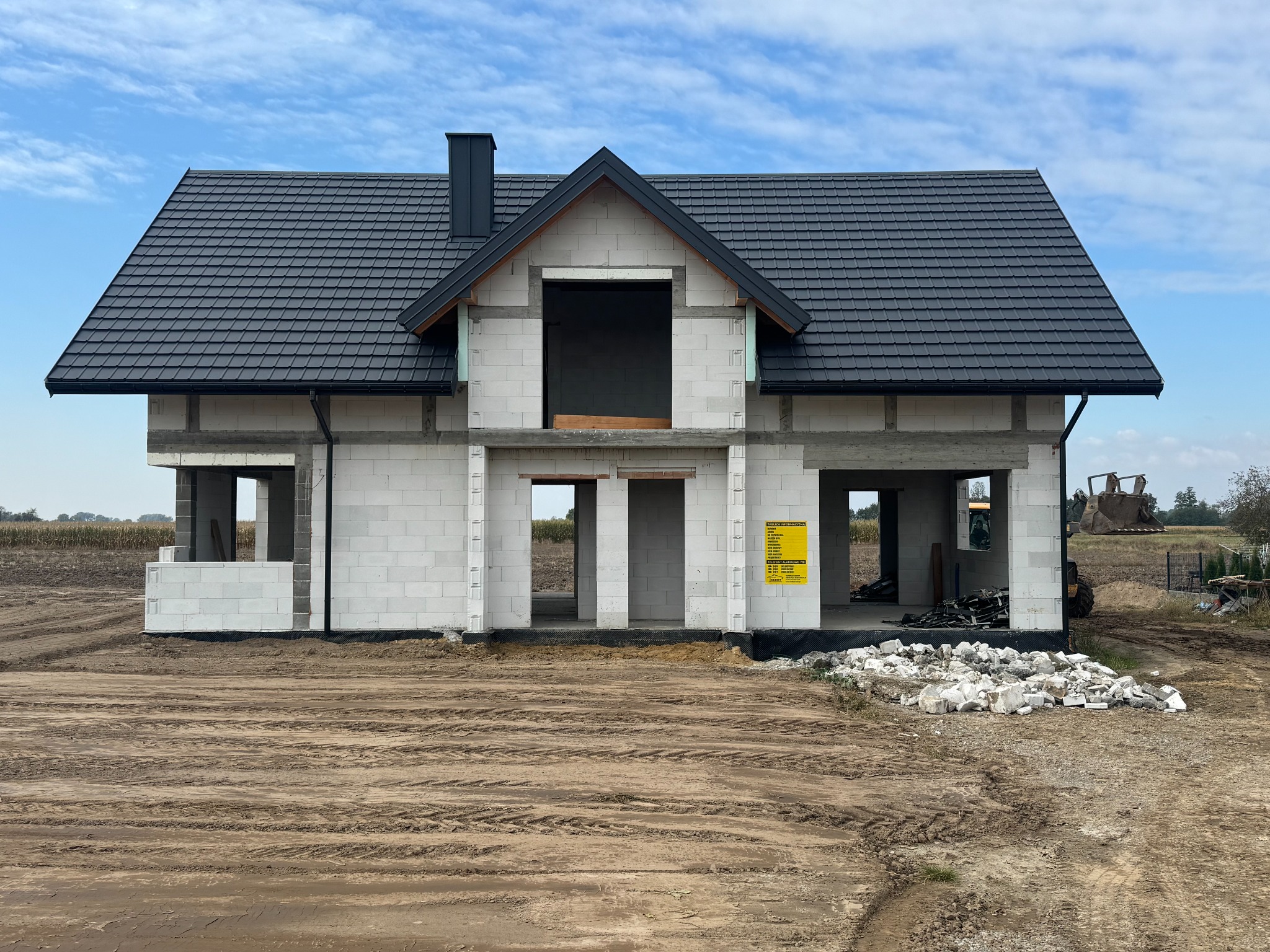 Surowy stan domu murowanego z ciemną dachówką, widoczne bloczki, otwory okienne i drzwiowe, teren budowy z ziemią i gruzem. W tle pole i błękitne niebo.