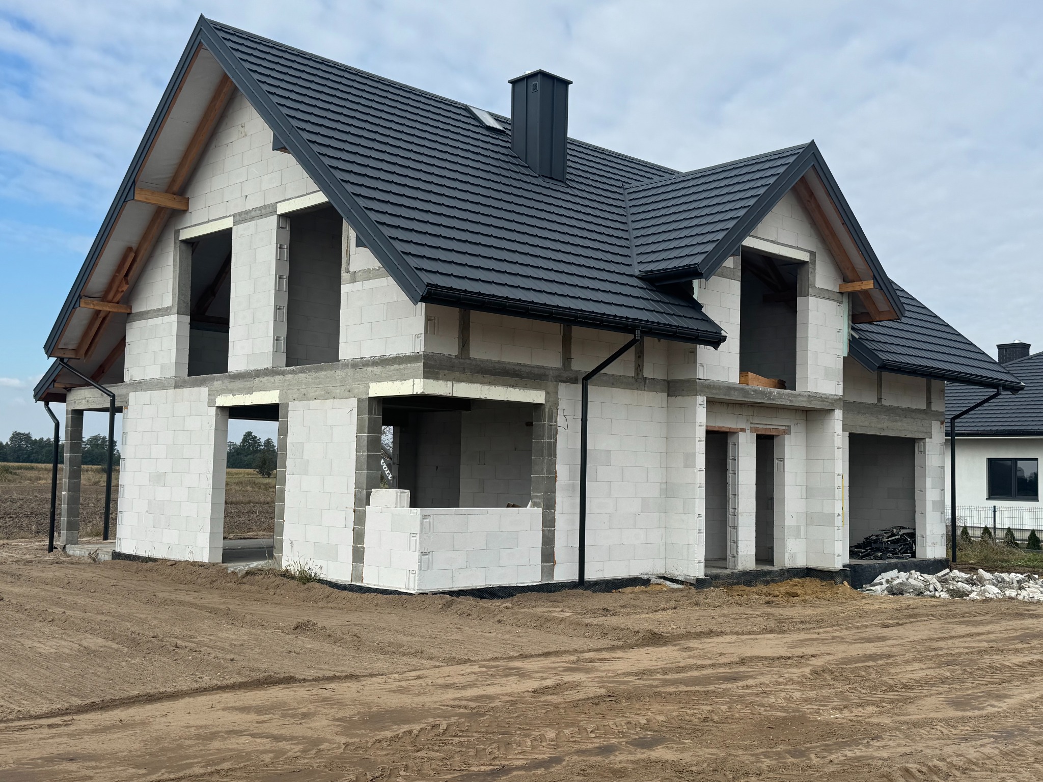 Surowy stan domu murowanego z czarną dachówką w Dąbrowie Tarnowskiej. Widoczne białe bloczki, brak okien i drzwi. Dom w trakcie budowy, teren wokół wyrównany.