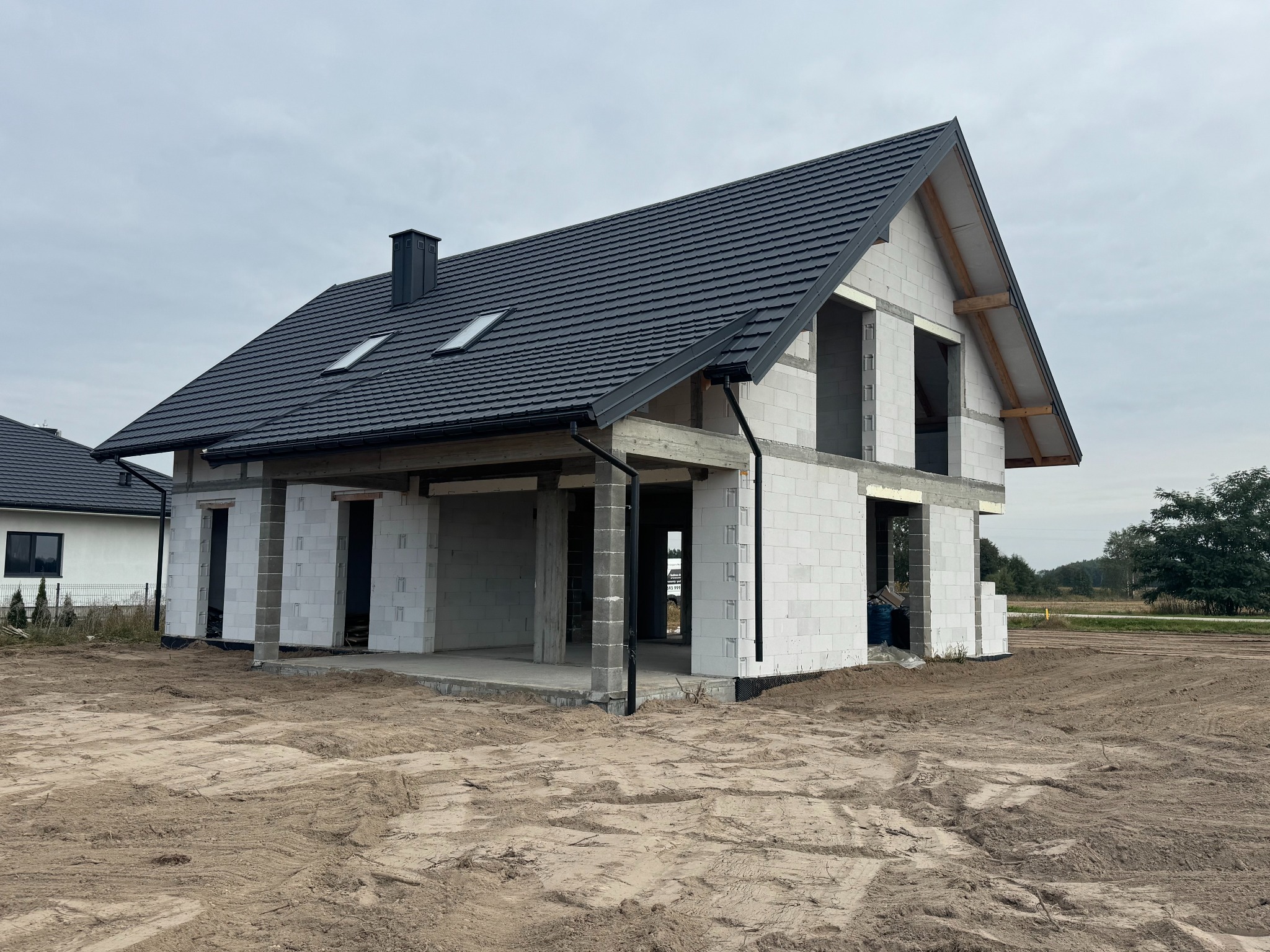 Surowy stan domu murowanego z czarną dachówką, widoczne okna dachowe i komin. Budynek w trakcie budowy, otoczony piaszczystym terenem. Widok z zewnątrz, w dzień.