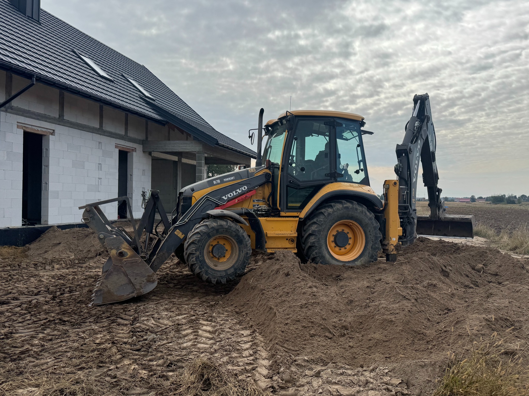 Żółta koparko-ładowarka Volvo na placu budowy domu z białego betonu komórkowego, z czarnym dachem i widocznymi oknami. Ślady opon na ziemi.