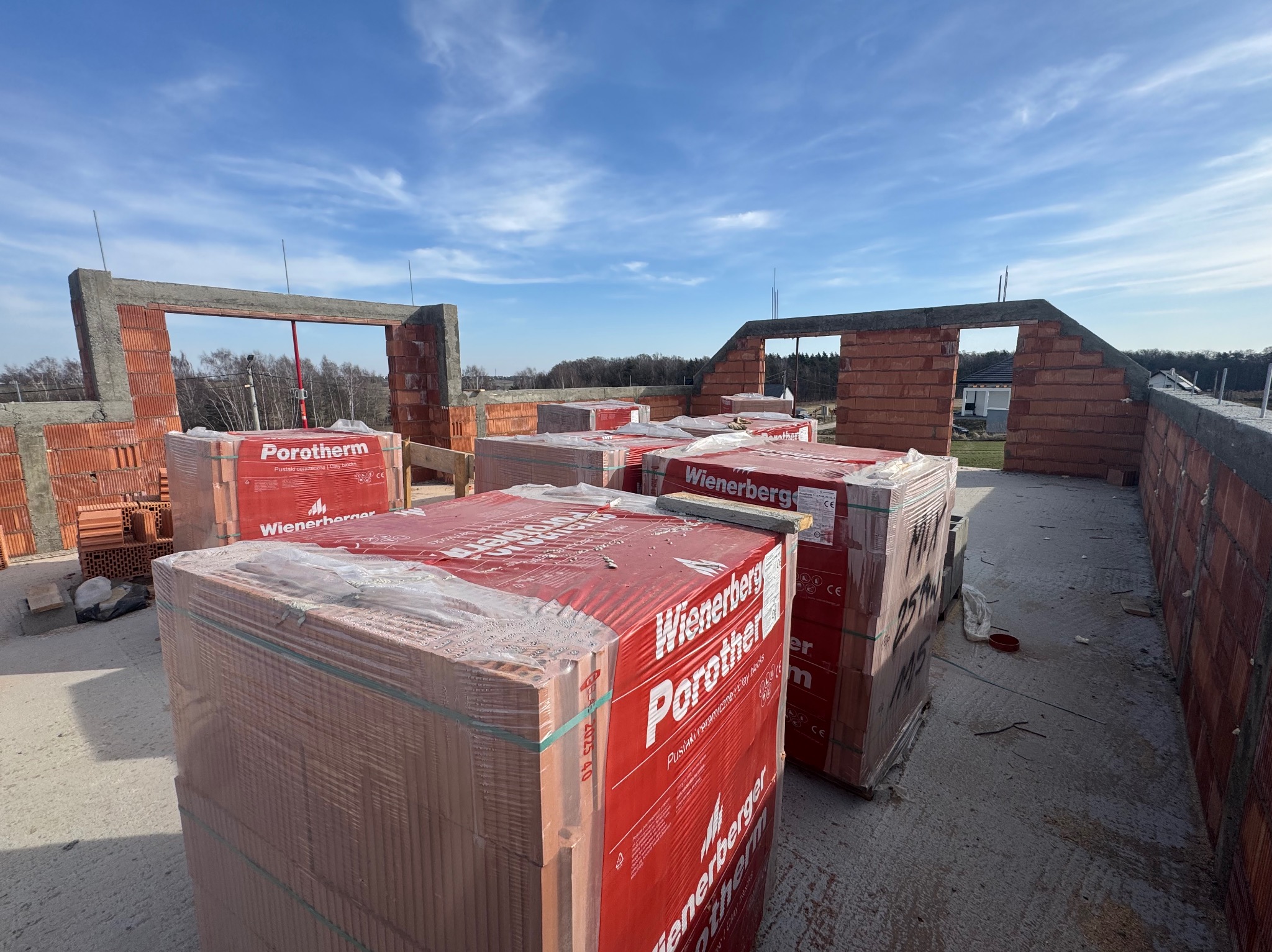 Budowa domu w toku: ściany z czerwonej cegły Porotherm, palety z pustakami ceramicznymi Wienerberger, błękitne niebo w tle. Widok z góry na plac budowy.