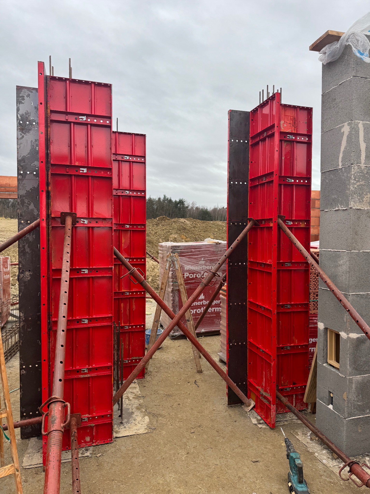 Czerwone szalunki ustawione pionowo, wsparte metalowymi stemplami, obok mur z bloczków betonowych. W tle palety z cegłami Porotherm.