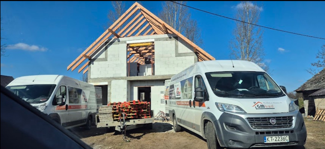 Budowa domu w stanie surowym otwartym z widocznym drewnianym szkieletem dachu. Na pierwszym planie dwa busy budowlane i przyczepka z elementami szalunkowymi.