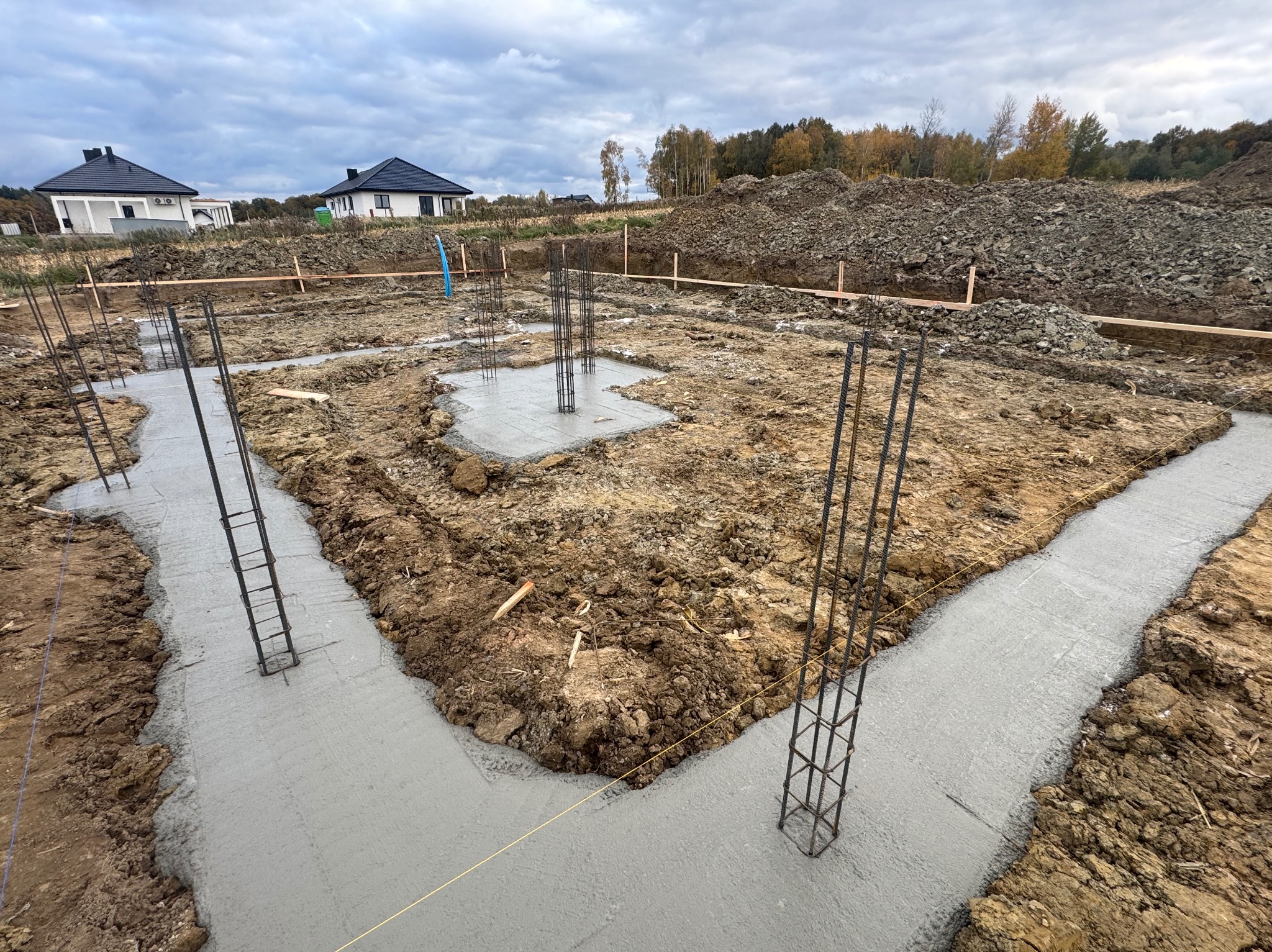 Wylane fundamenty pod budowę domu jednorodzinnego. Widoczne zbrojenia i świeży beton w wykopach. W tle dwa domy i hałda ziemi pod pochmurnym niebem.