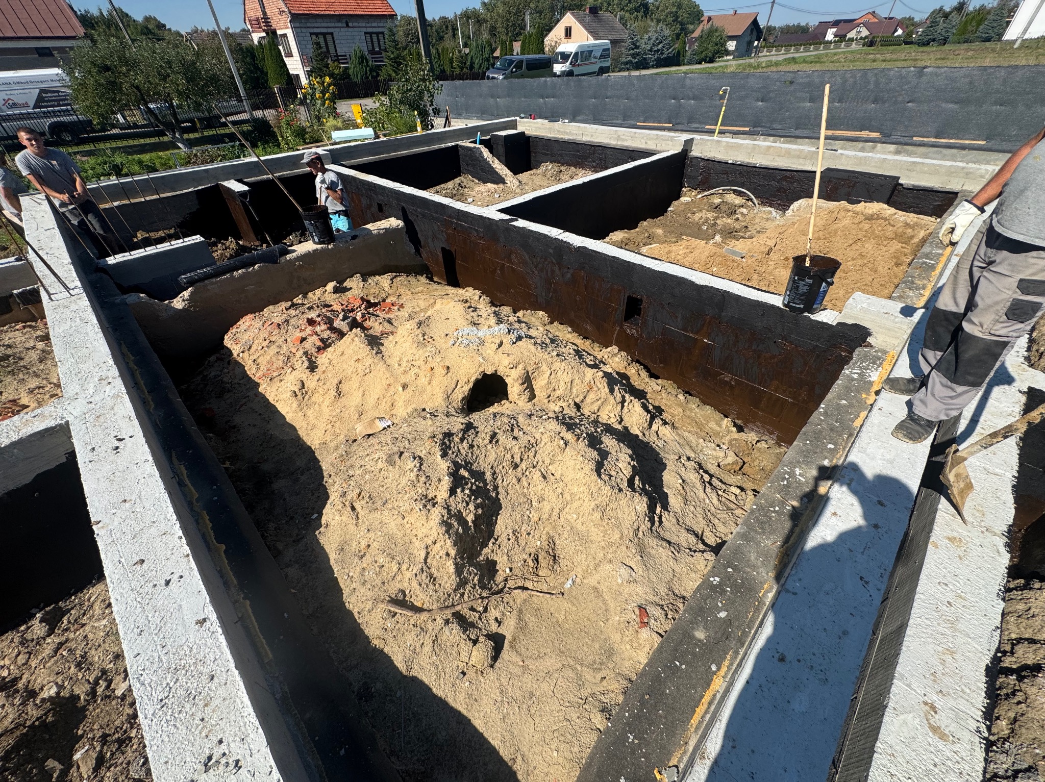 Fundamenty z izolacją bitumiczną w Dąbrowie Tarnowskiej. Wnętrze wypełnione piaskiem, widoczni pracownicy budowlani. Ujęcie z góry, w tle domy jednorodzinne.
